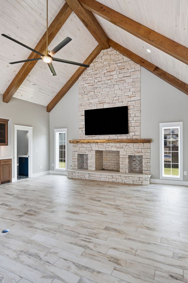 Spacious living room with whitewashed floors, stone fireplace, vaulted wood beams, and wall-mounted TV