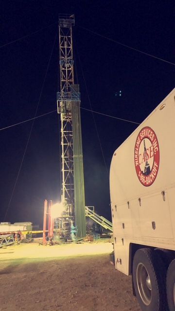 Oil drilling rig lit up at night, with a white truck bearing a company logo in the foreground.