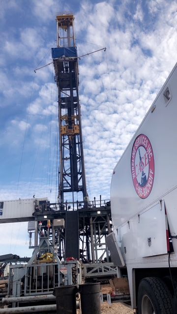 Drilling rig against a cloudy sky, next to a white service truck with logo.
