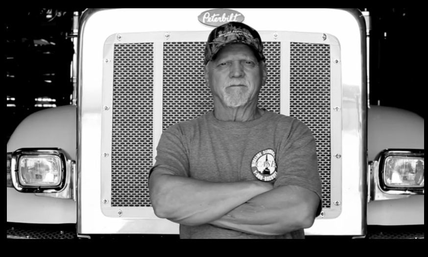 Man with crossed arms in front of a Peterbilt truck, wearing a camouflage hat and t-shirt.
