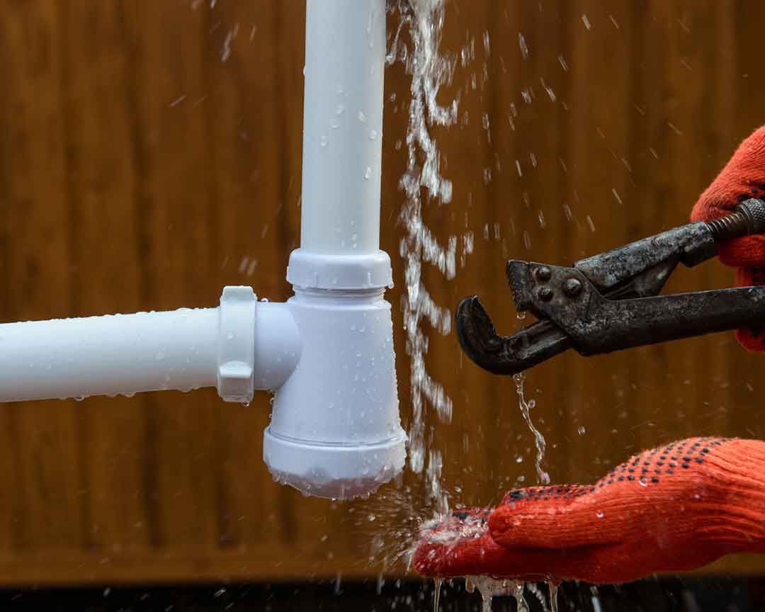 Plumber in orange gloves using wrench to fix a leaking white PVC pipe.