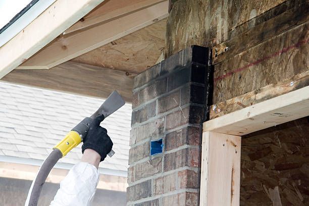 Person using a tool to clean bricks on a building's exterior with visible wood framing and a portion of a roof.