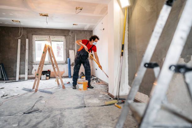 Person painting a room, wearing a mask and work overalls; ladders and painting supplies present.