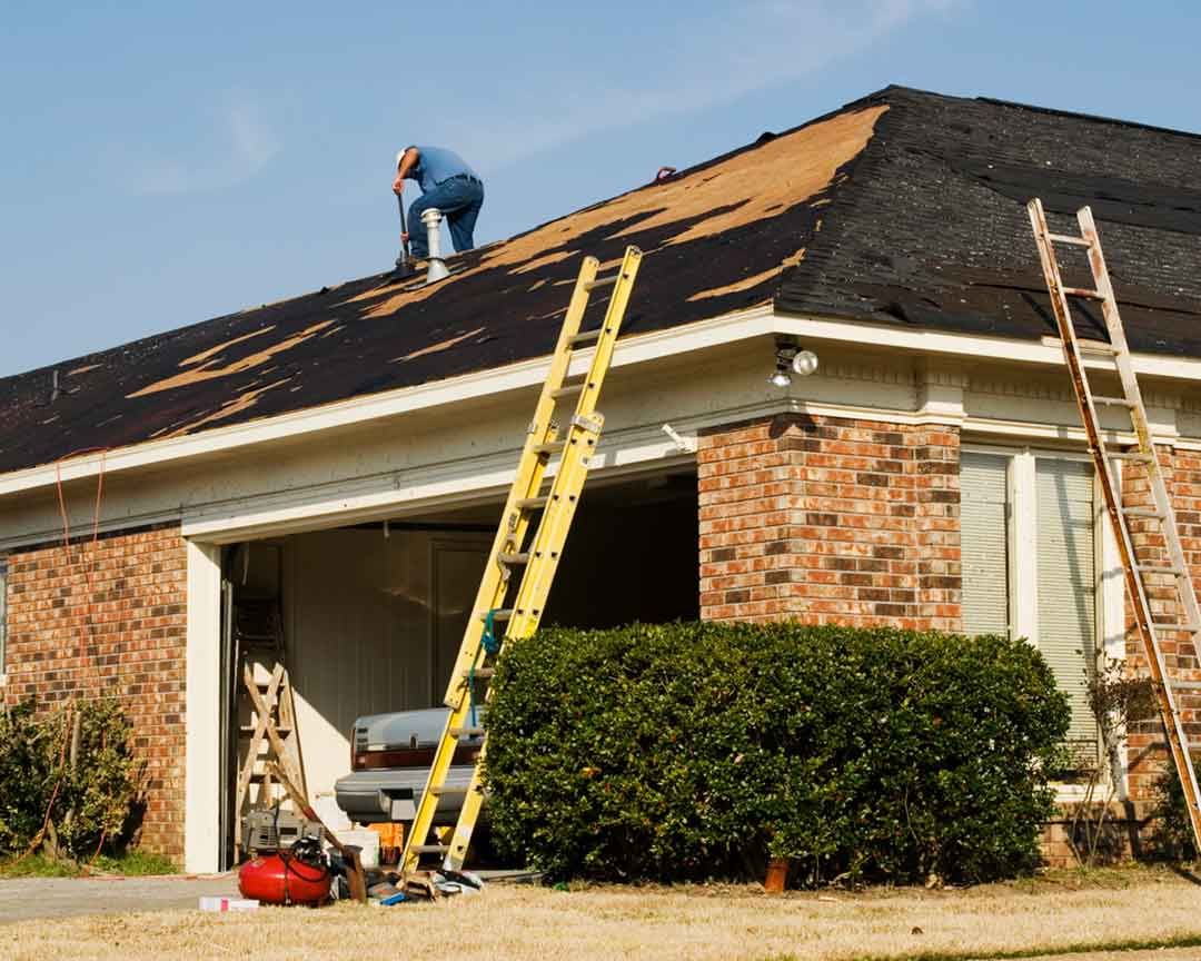 Roofer on a house roof removing shingles with ladders leaning against the building.