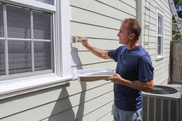 Man painting house siding, using brush and paint tray. Outdoors, next to a window.