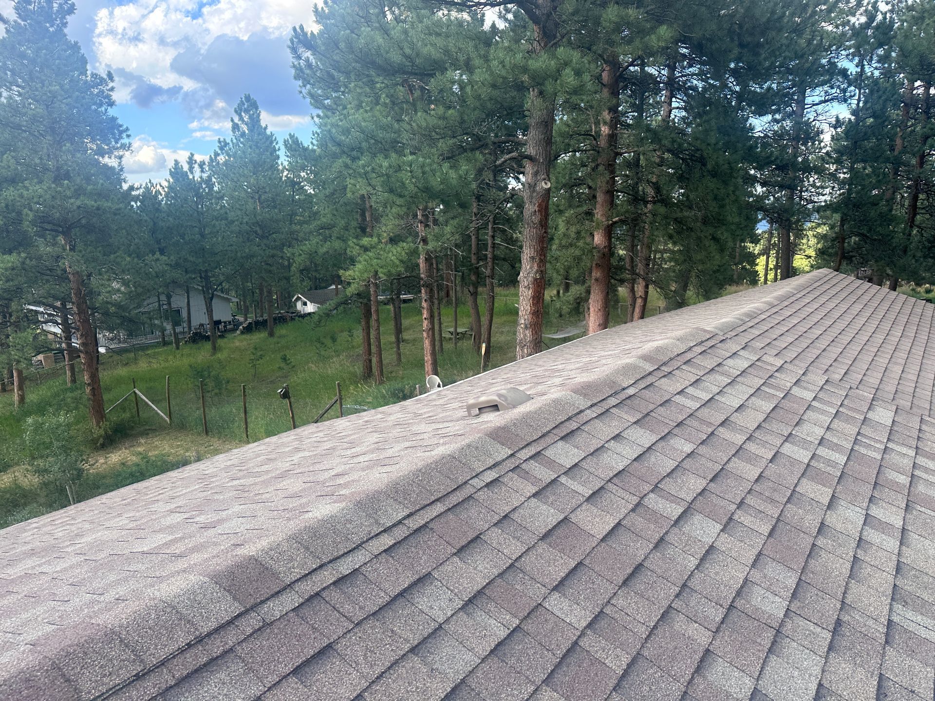 Roof with brown shingles, trees, and green hillside in the background under a partly cloudy sky.