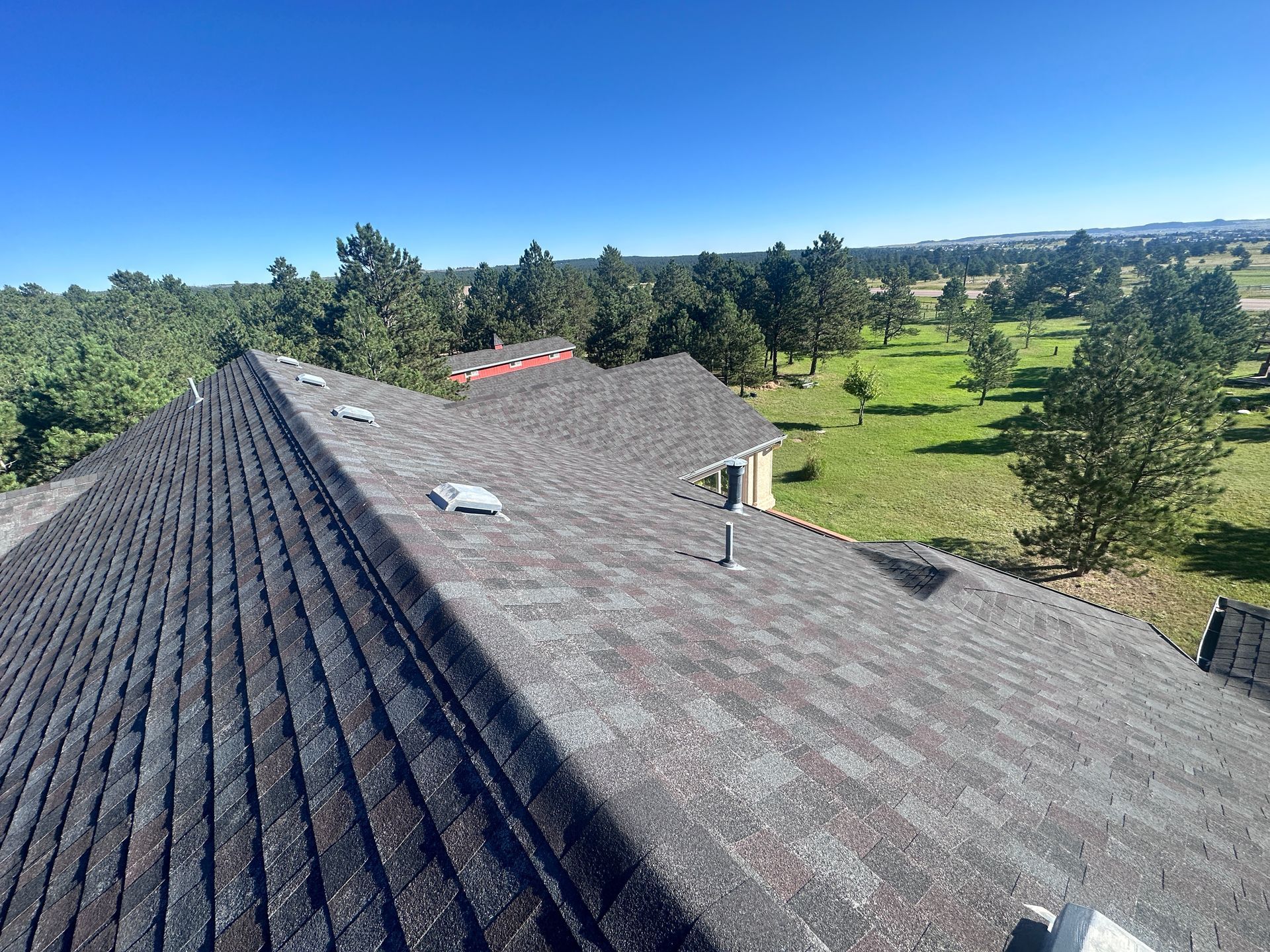 View from a house roof overlooking a green field and trees on a sunny day. Gray shingles.