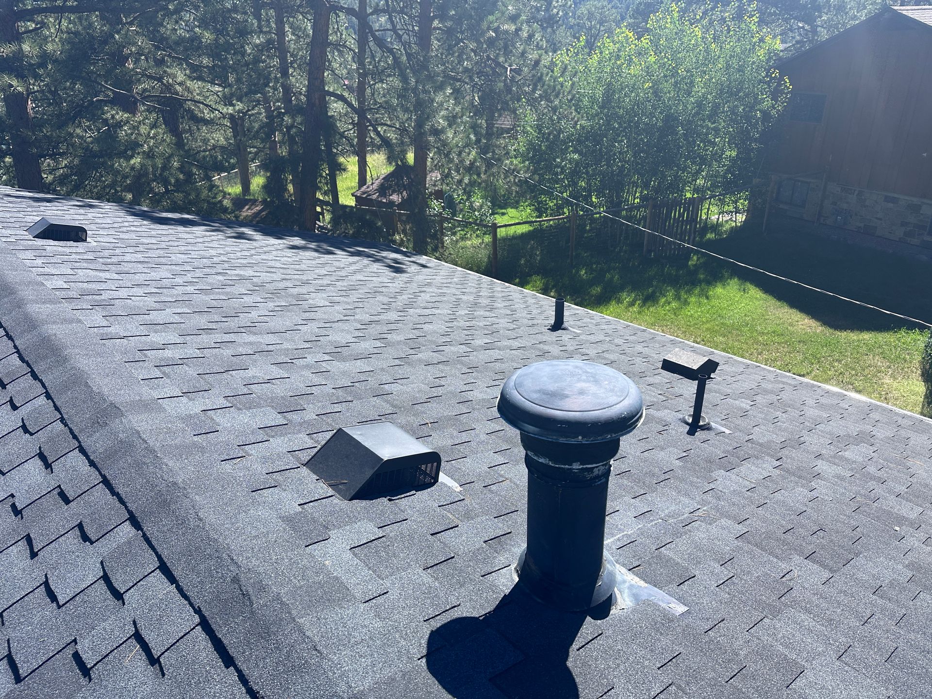 A dark-colored shingled roof with a vent, surrounded by trees under a sunny sky.