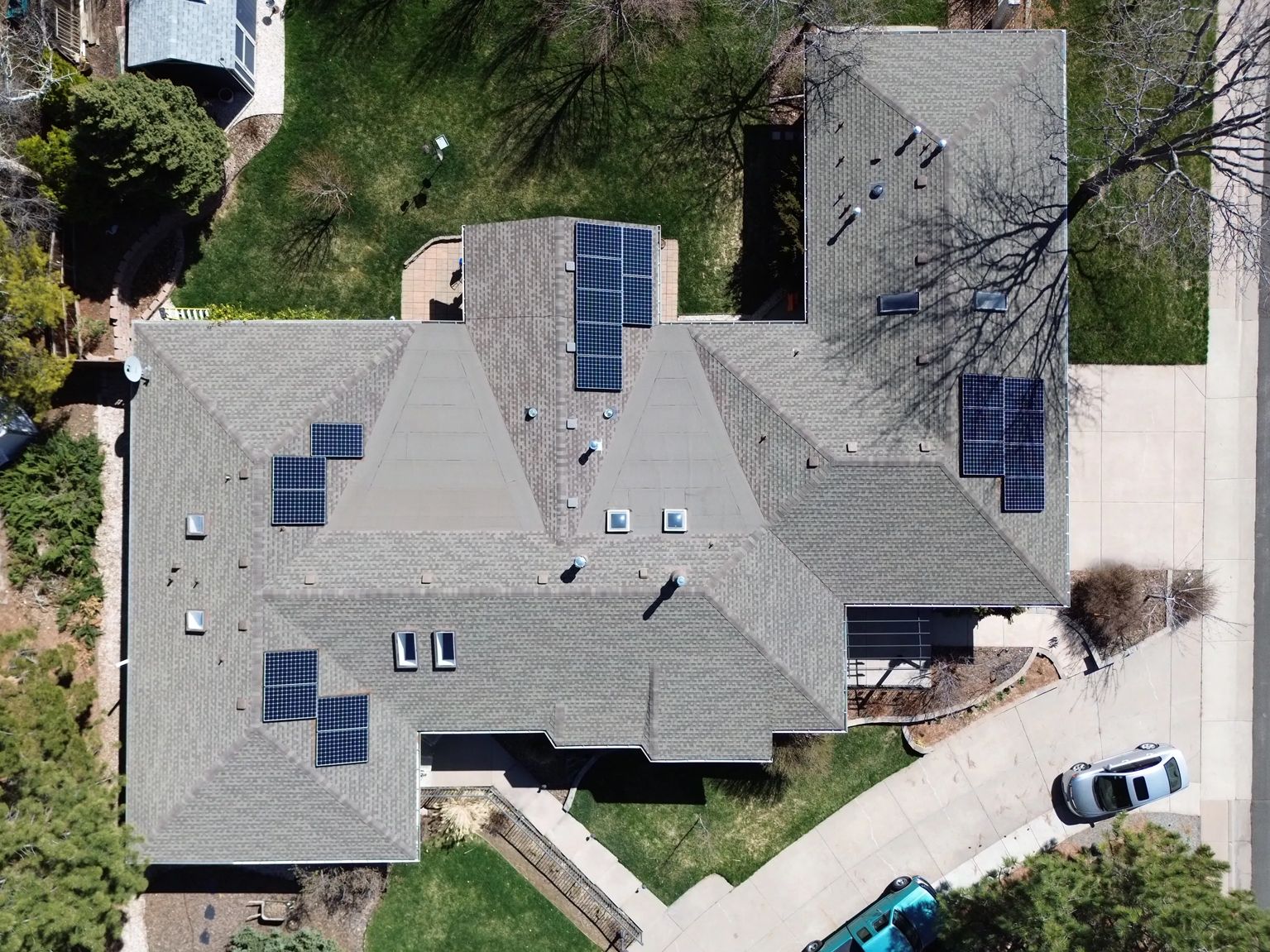 Overhead view of a house with solar panels on its gray shingled roof, a driveway, and surrounding greenery.