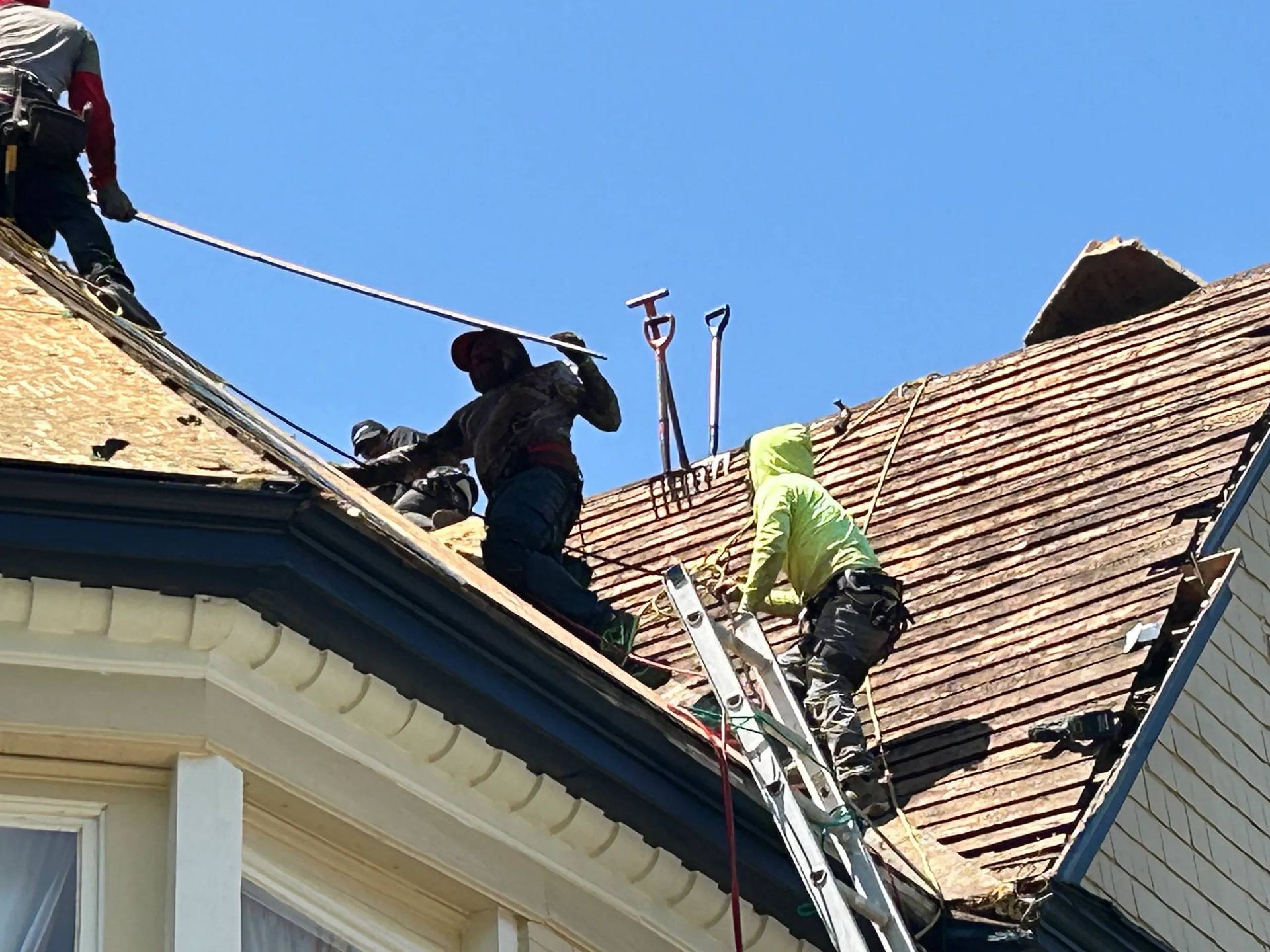 Roofers working on a house under a clear blue sky, using ropes, ladders, and tools to remove shingles.