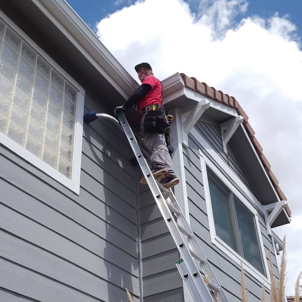 Man on a ladder cleaning a window on a gray house under a cloudy sky.