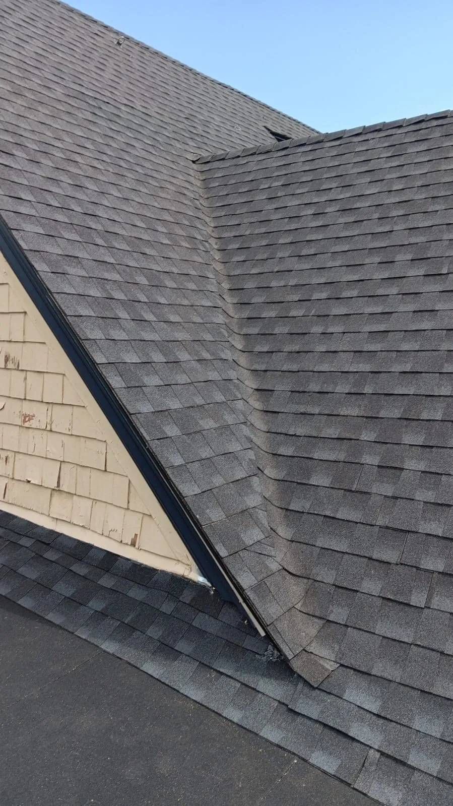 Gray asphalt shingle roof with a light-colored building wall against a clear blue sky.