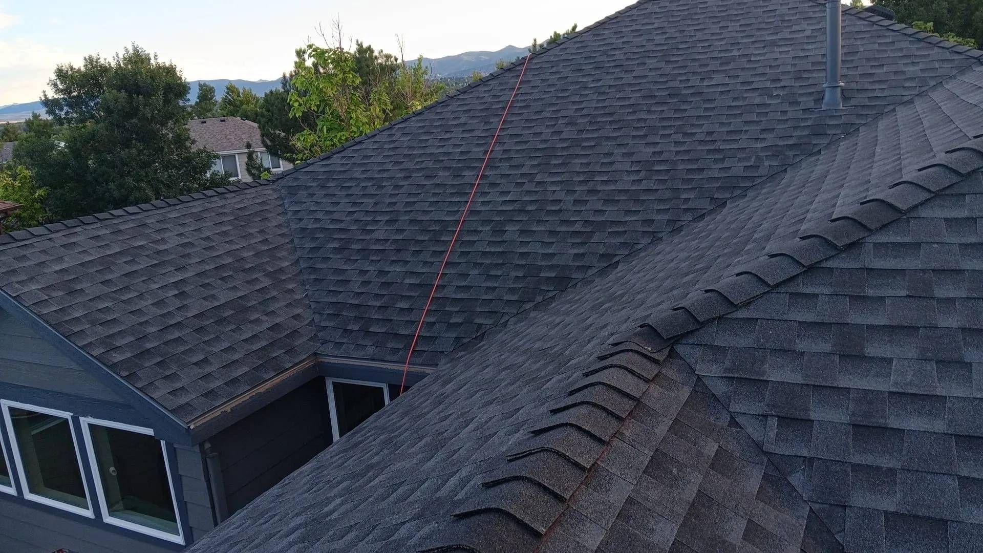 Dark gray asphalt shingle roof, aerial view. House with windows, trees, and mountains in background.