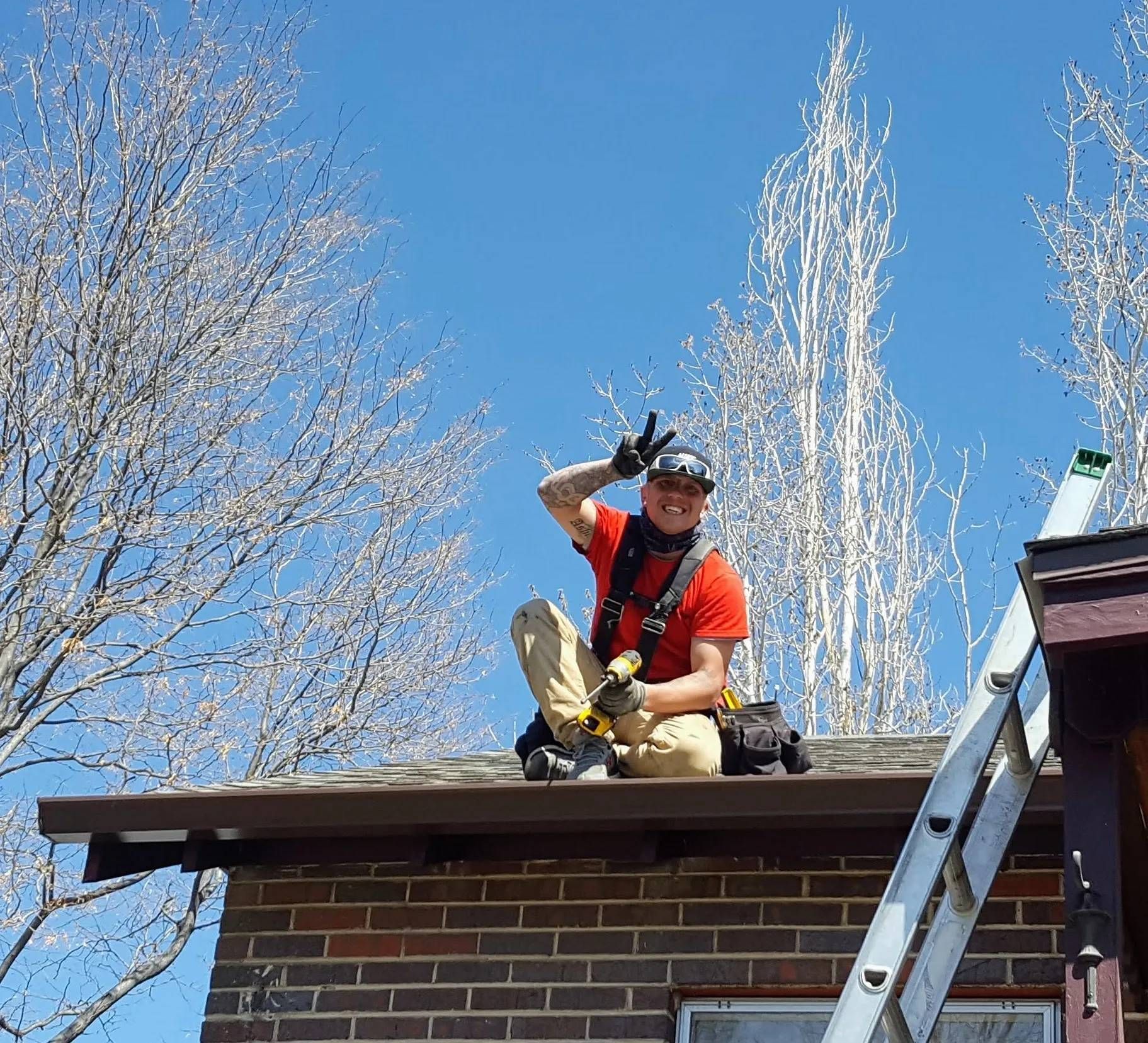 Person on a roof gives a peace sign, wearing a safety harness. Blue sky, brick building, bare trees.