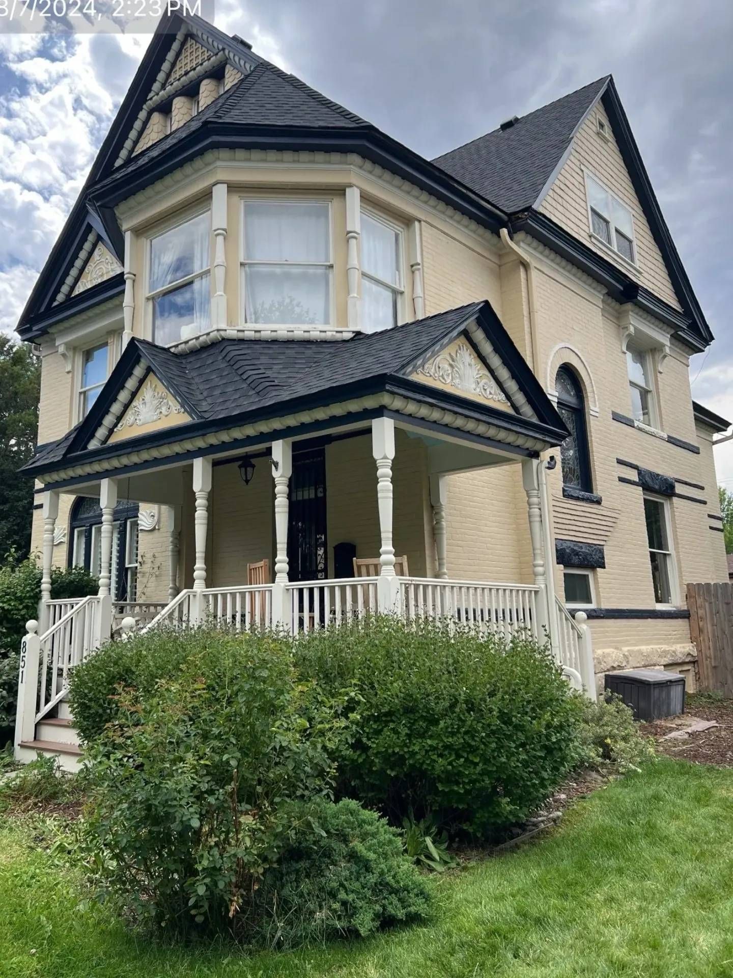 Victorian-style house with yellow siding, black roof, and a porch. Green bushes and grass in front.