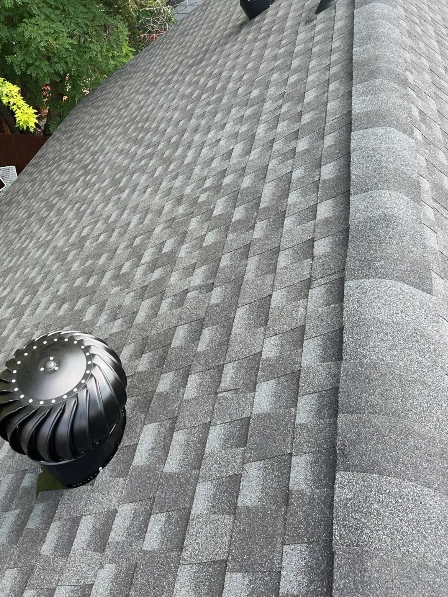 Gray asphalt shingle roof with black turbine vent, viewed from above, surrounded by green trees.