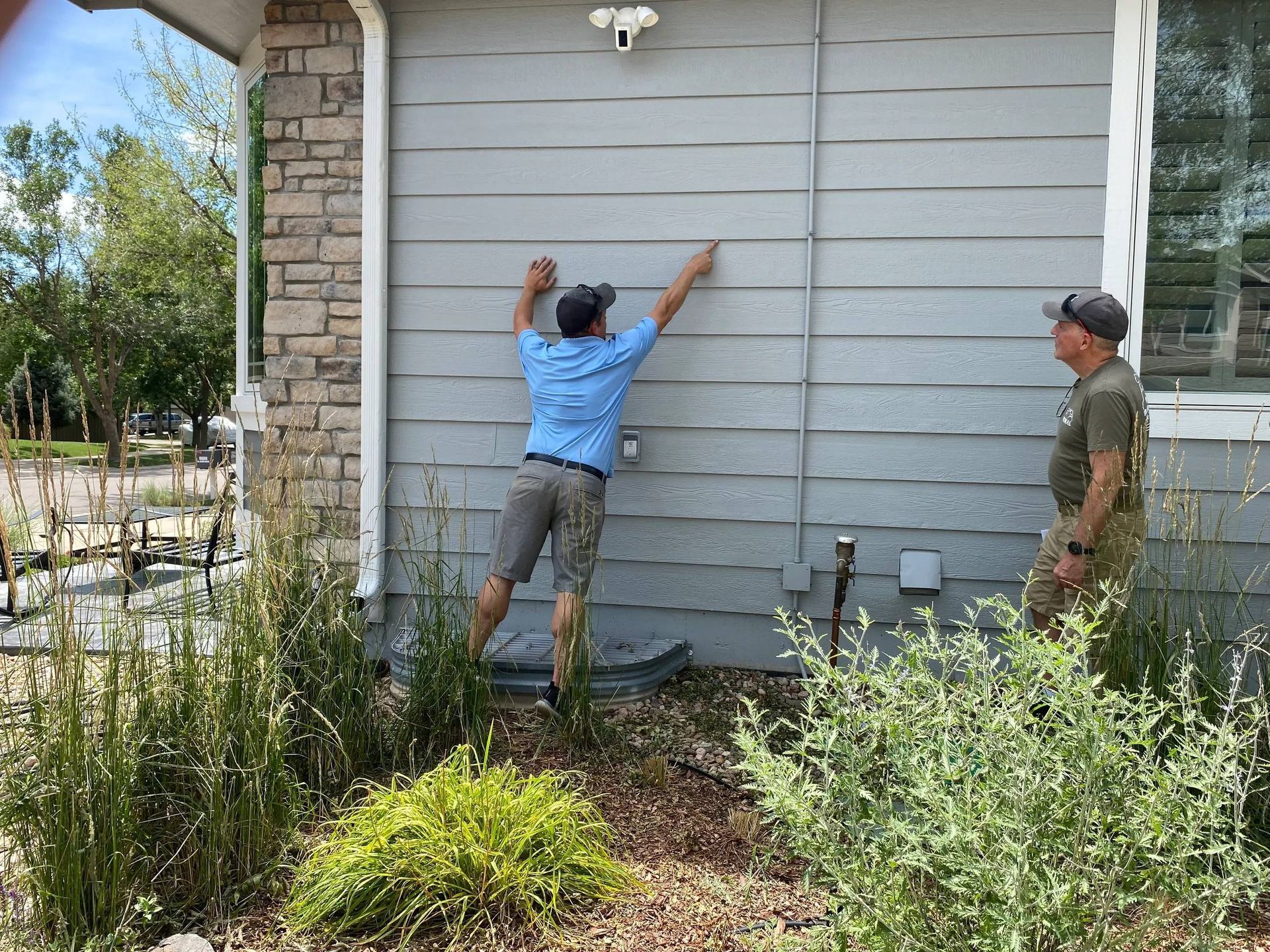 Two men installing a security camera on a light gray house wall, surrounded by plants.