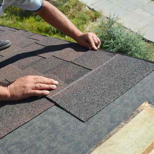 Person installing asphalt shingles on a roof, hands in view.