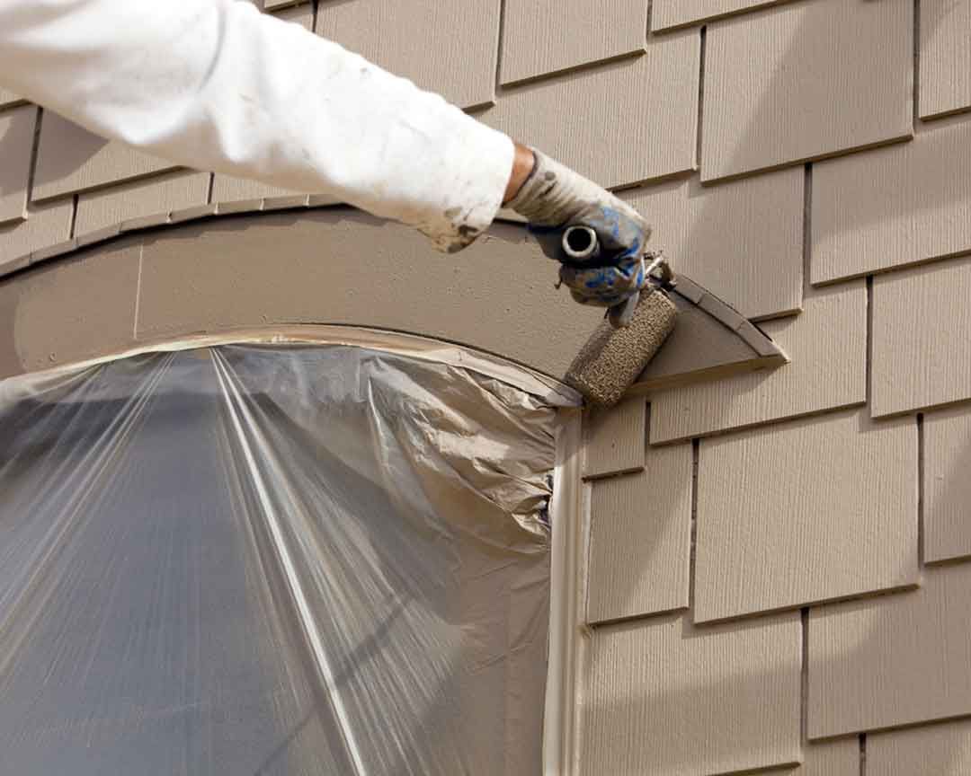 Person painting a window frame with a paint sprayer; window covered in plastic, cedar shake siding.