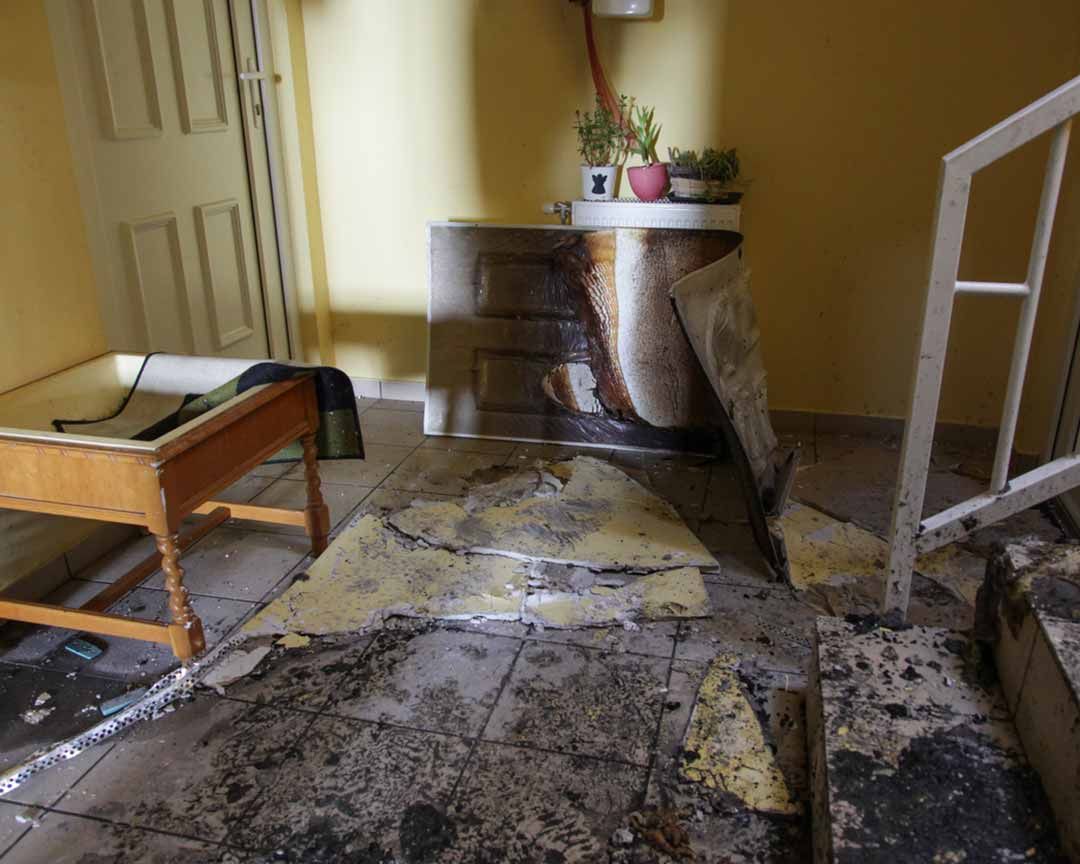 Damaged interior with peeling paint, debris on floor. A small table and a plant stand are visible.