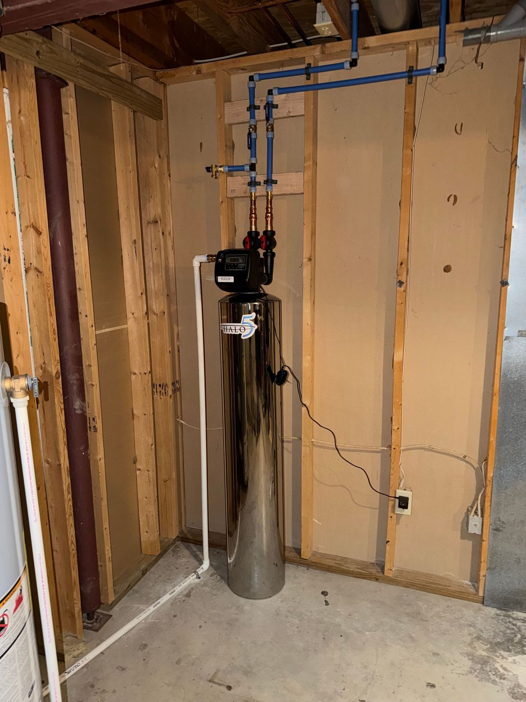 Water softener tank and control valve installed in an unfinished basement with exposed studs.