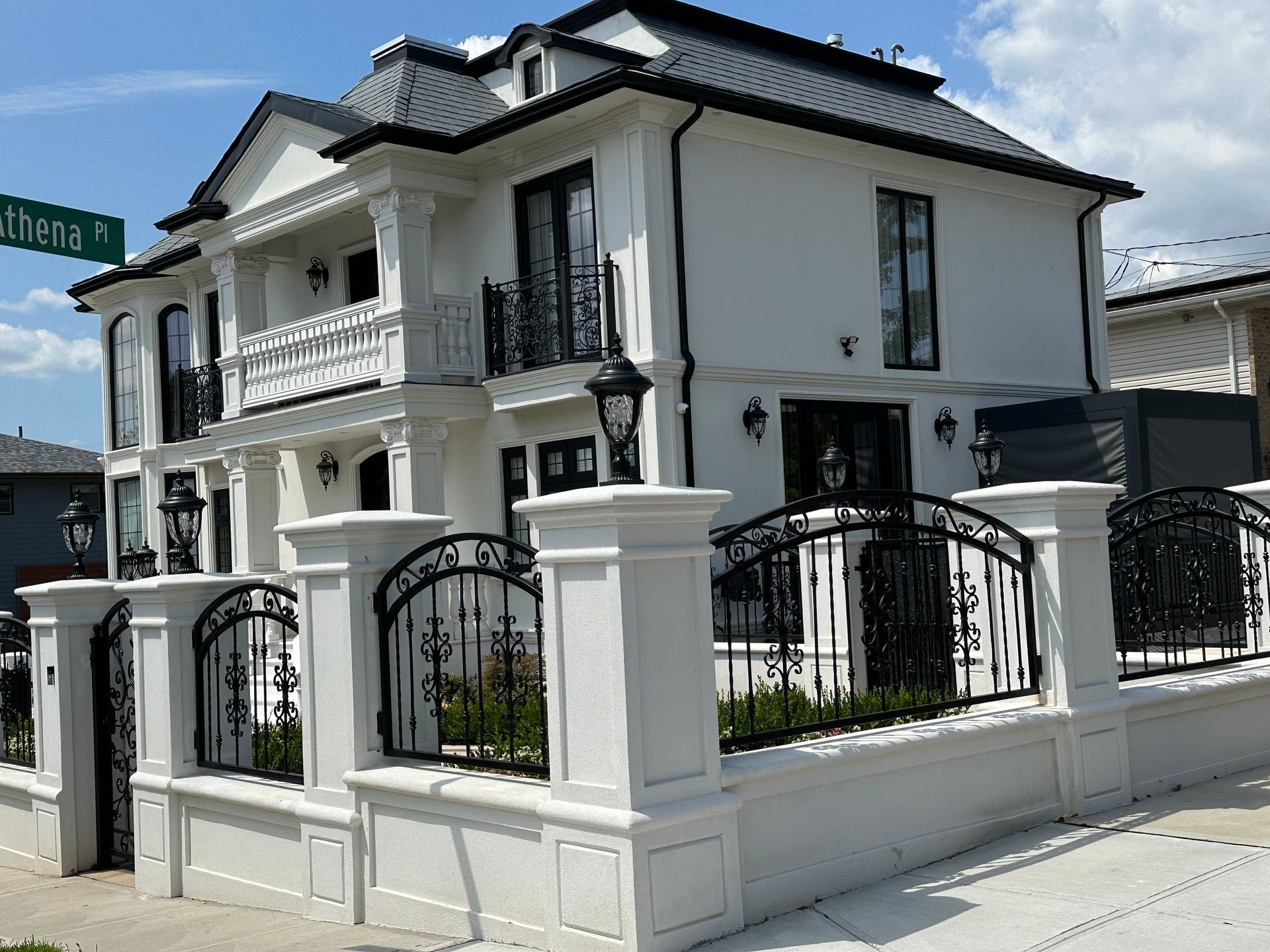 Modern white two-story house with black roof, balcony, and wrought-iron fence along the street