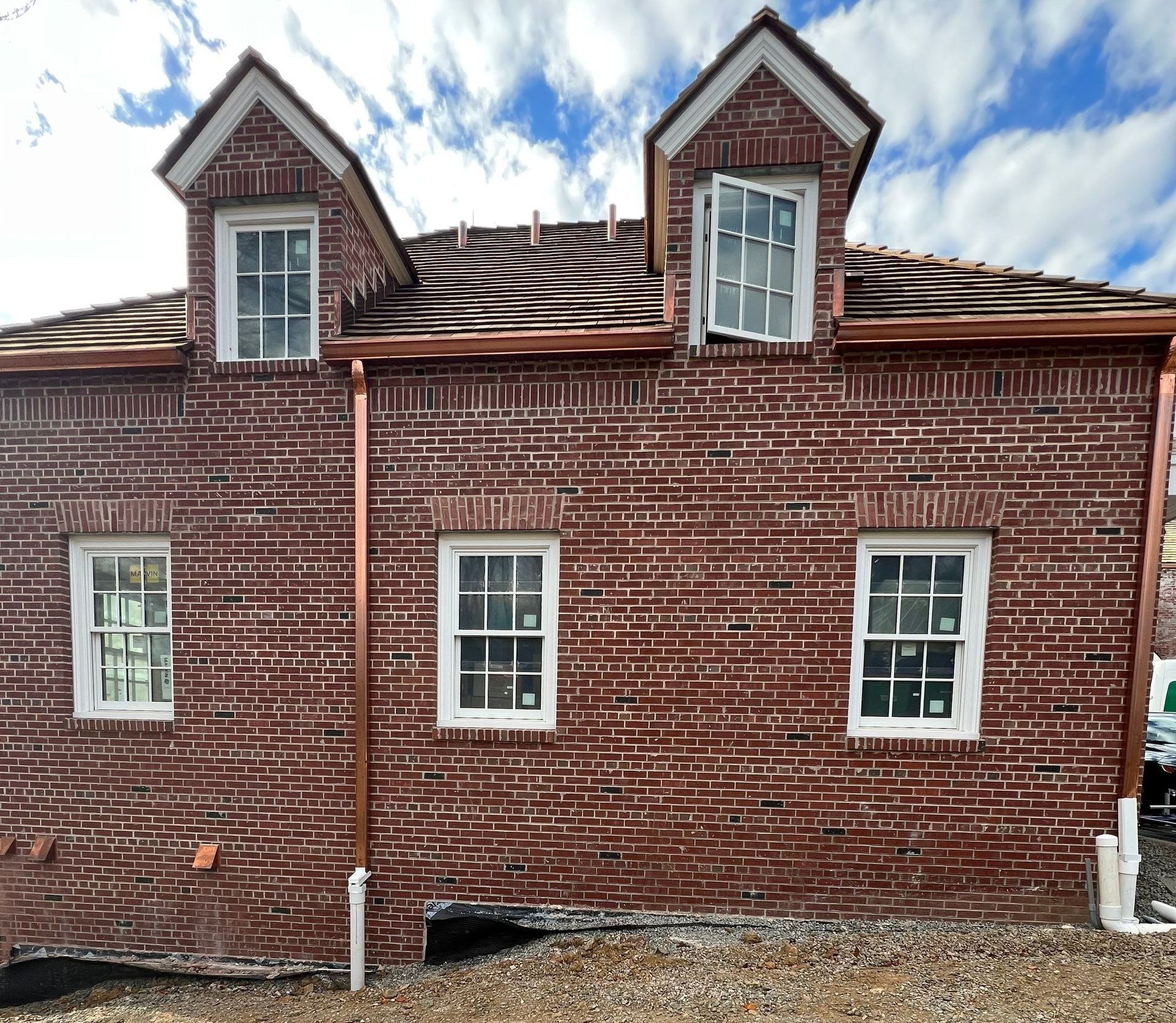 Red brick house side wall with white-trimmed windows and two dormer windows under a damaged roof.