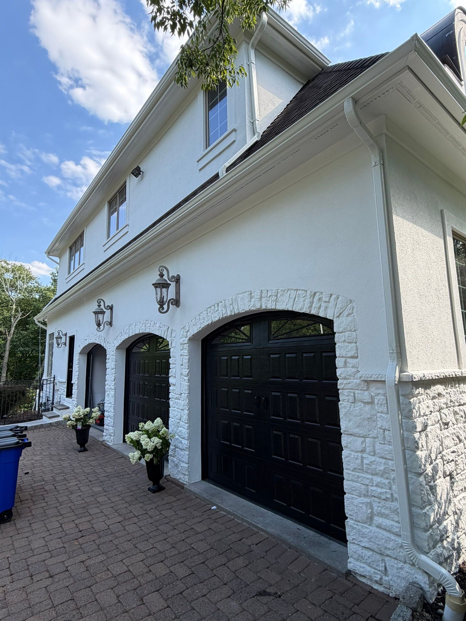 White two-story house with black garage doors, stone accents, and a brick driveway under a blue sky.