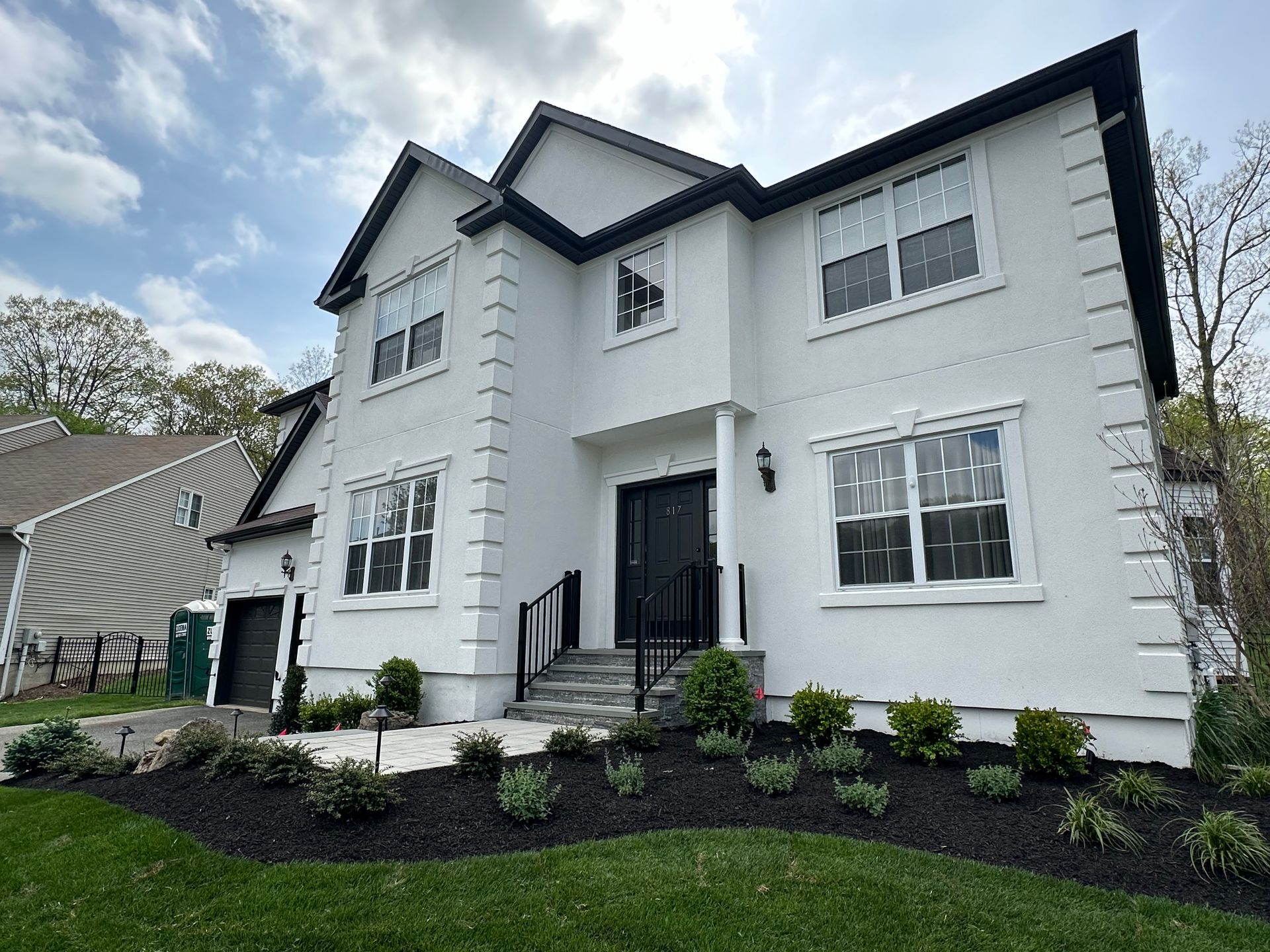 Large white two-story house with black trim, front steps, and landscaped yard under a cloudy sky.