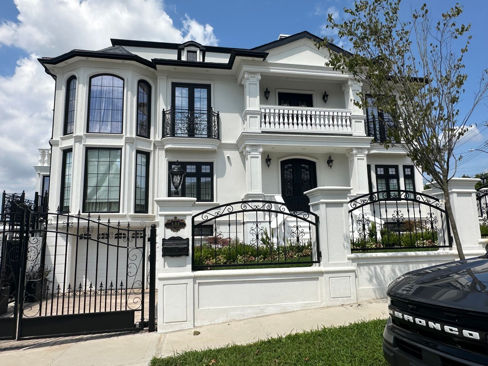 White two-story luxury house with black trim, balcony, and wrought-iron fence under a blue sky.