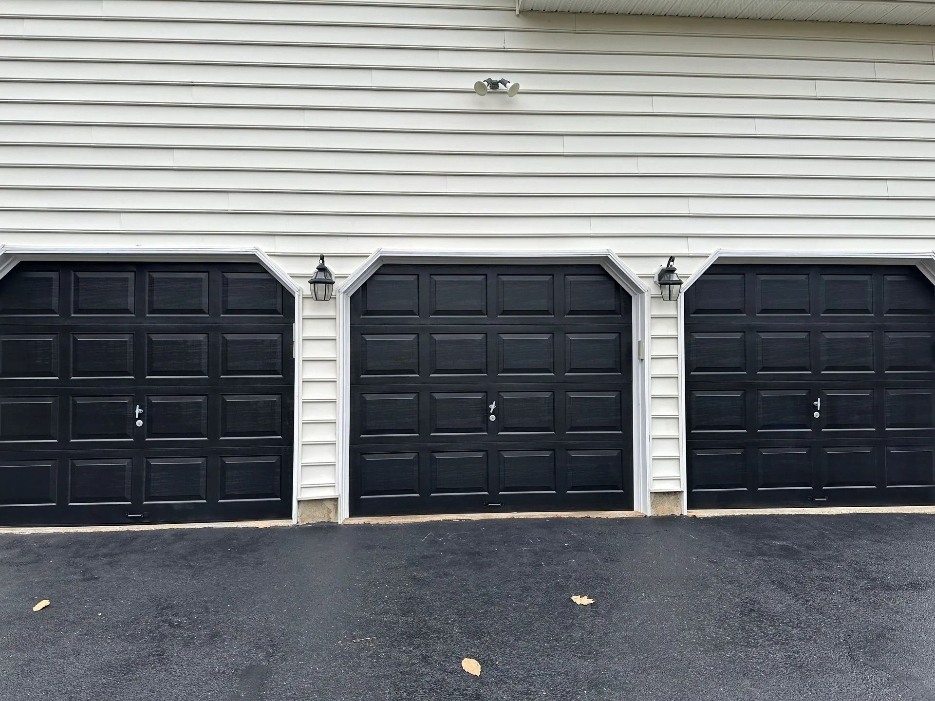 Three black garage doors on a white-sided house with a dark driveway.