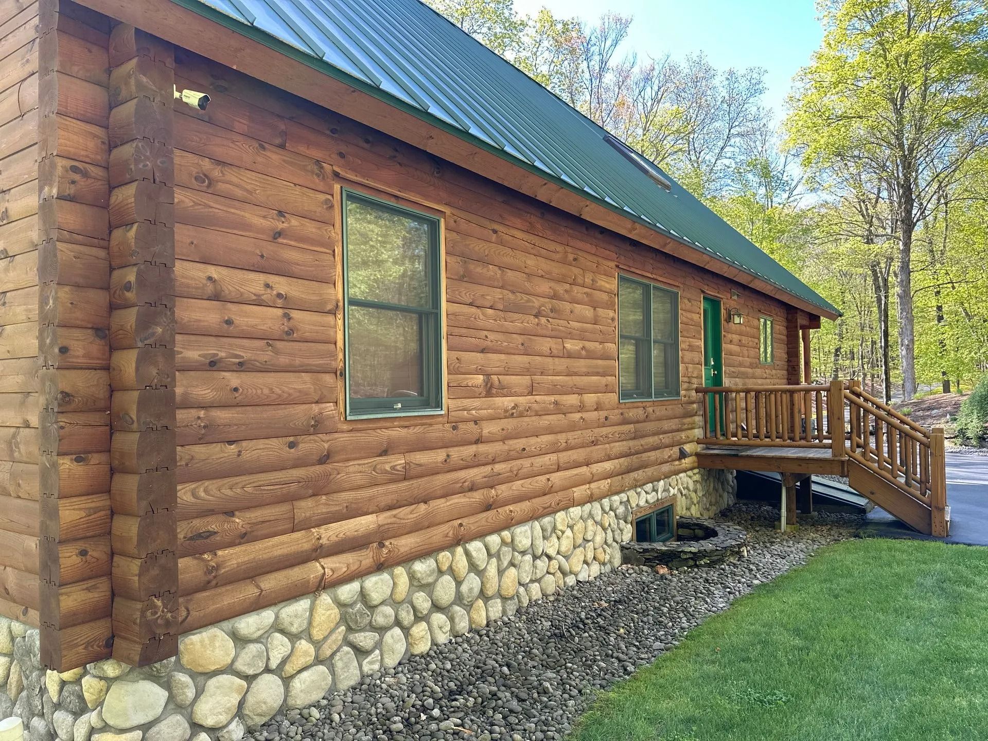 Log cabin with stone foundation, green roof, and wooden porch in a grassy yard.