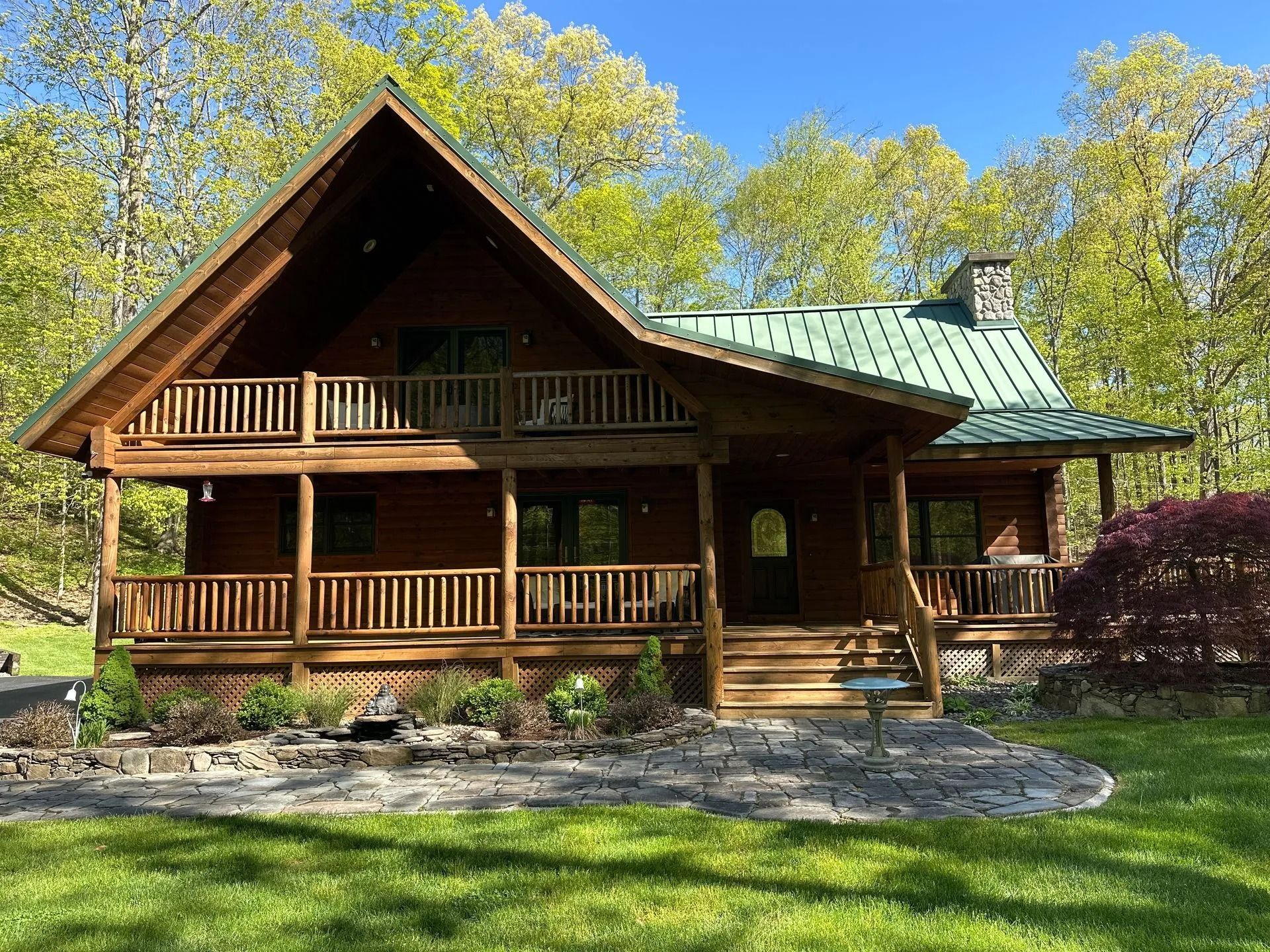 Rustic log cabin with green metal roof and wraparound porch in a sunny wooded clearing