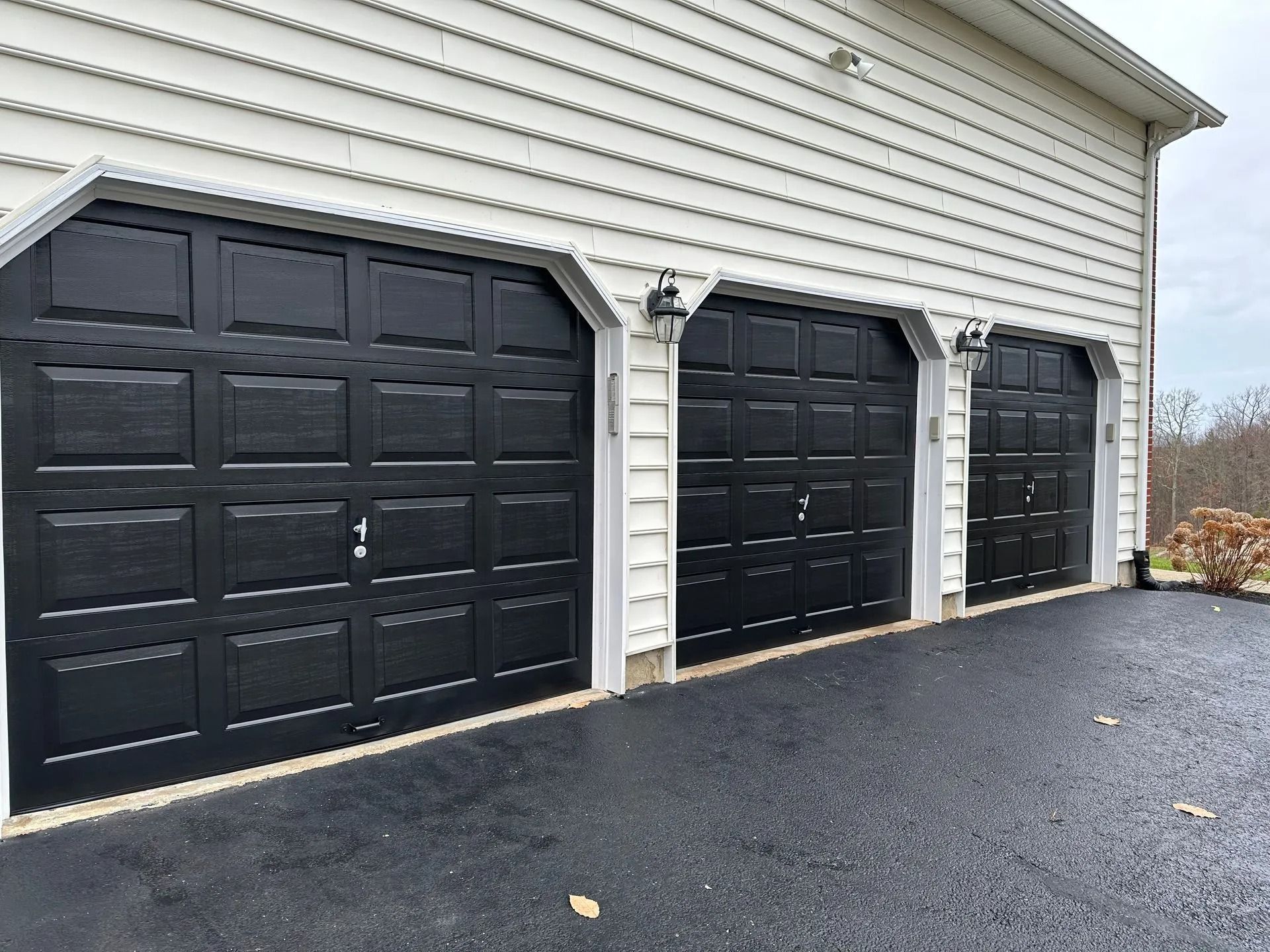 Three black garage doors on a white-sided house beside a paved driveway
