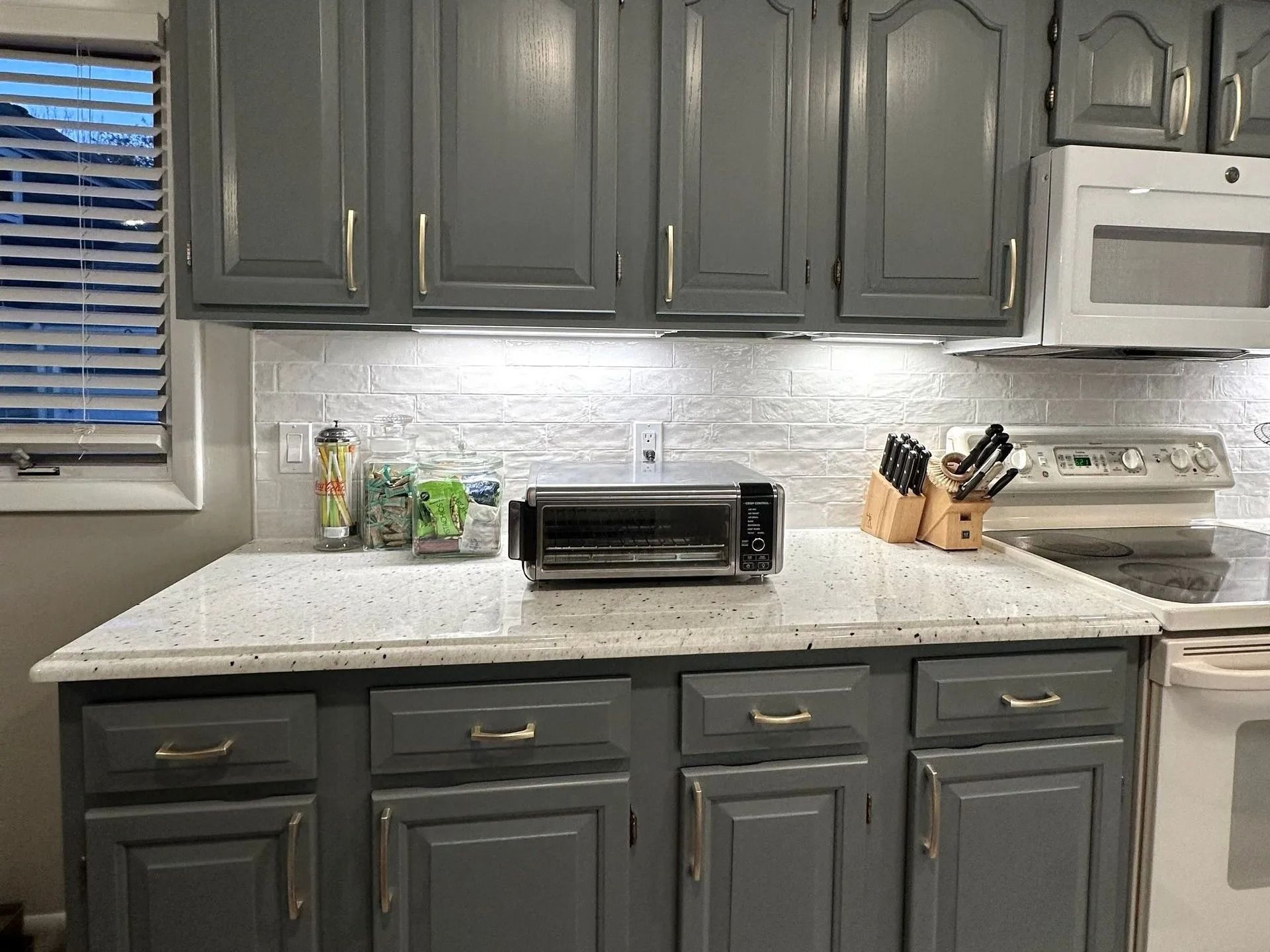 Gray kitchen cabinets with white countertop, toaster oven, knife block, and white stove under window blinds.