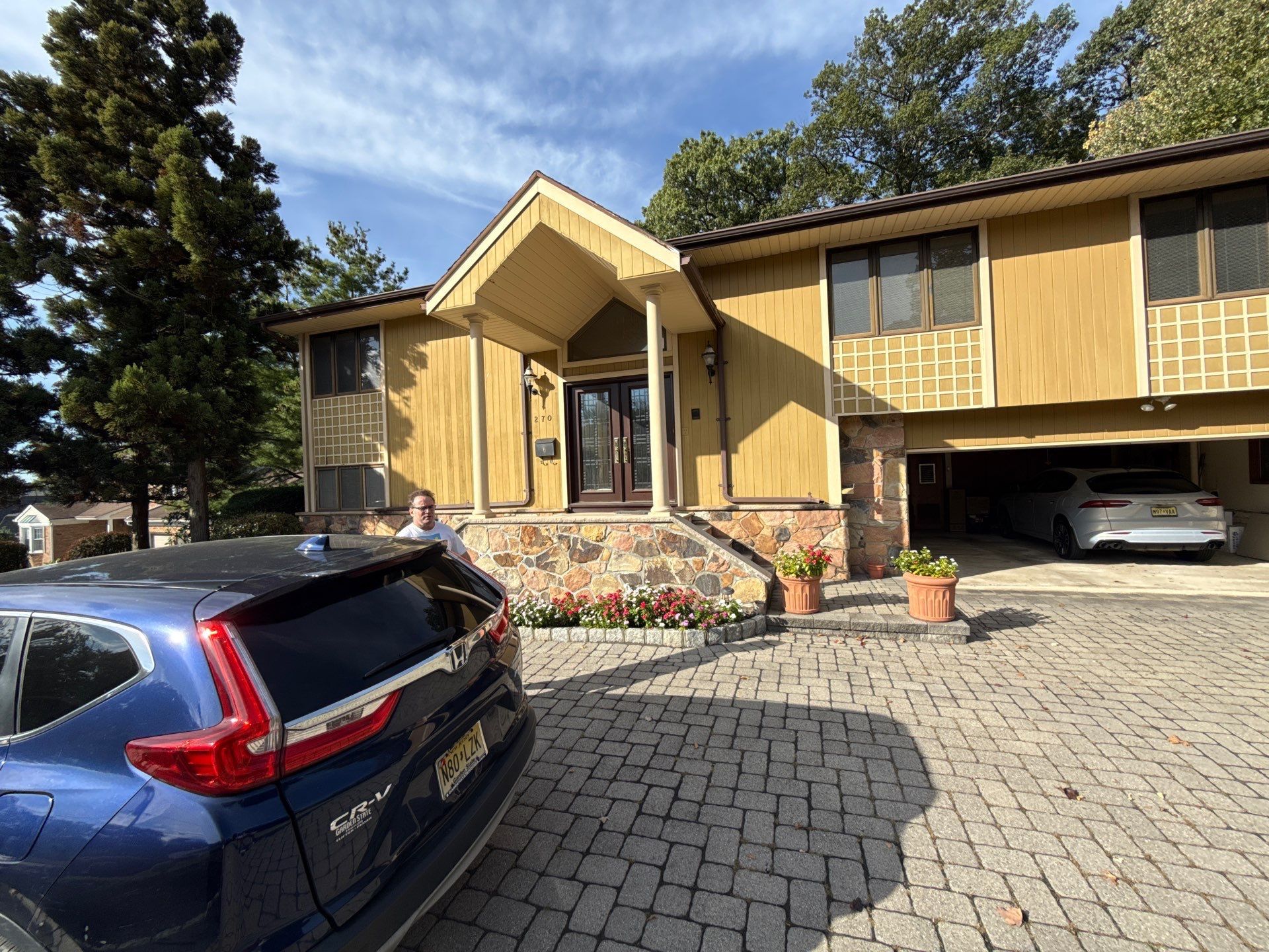 Suburban house with a covered entry, stone steps, driveway, and parked cars under a blue sky
