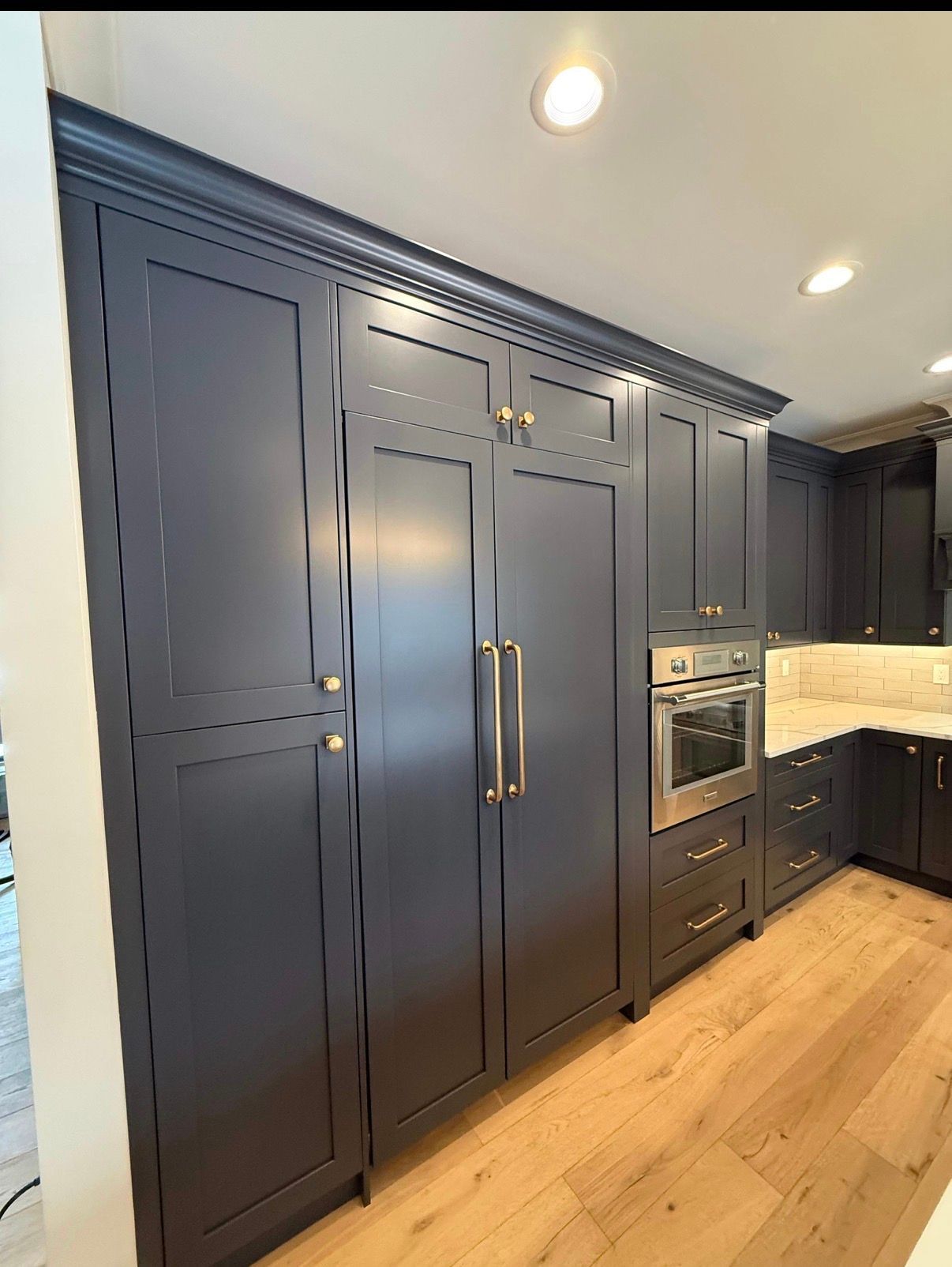 Dark blue kitchen cabinets with built-in oven, brass handles, and light wood floors under recessed lights
