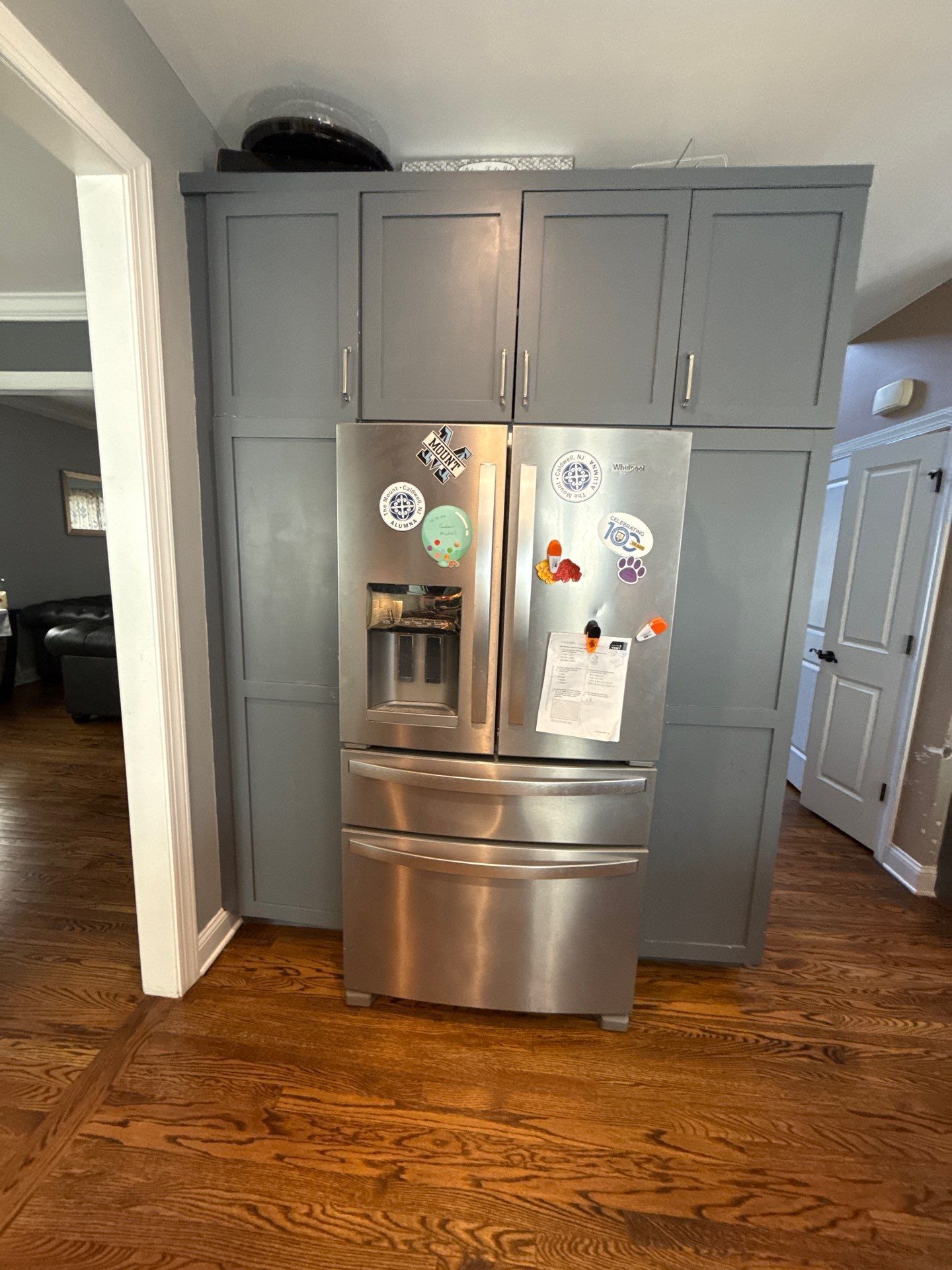 Stainless steel refrigerator with magnets and papers, built into gray kitchen cabinets on a hardwood floor.