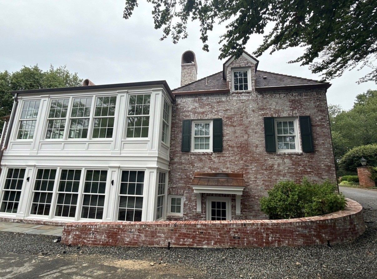 Brick house with white sunroom and dark shutters, surrounded by stone wall and greenery