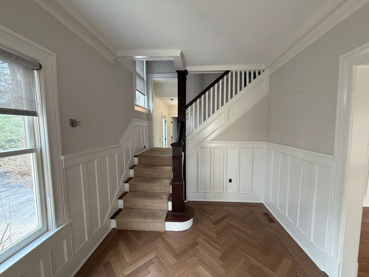Bright hallway with herringbone wood floor, white wainscoting, and a staircase leading up.
