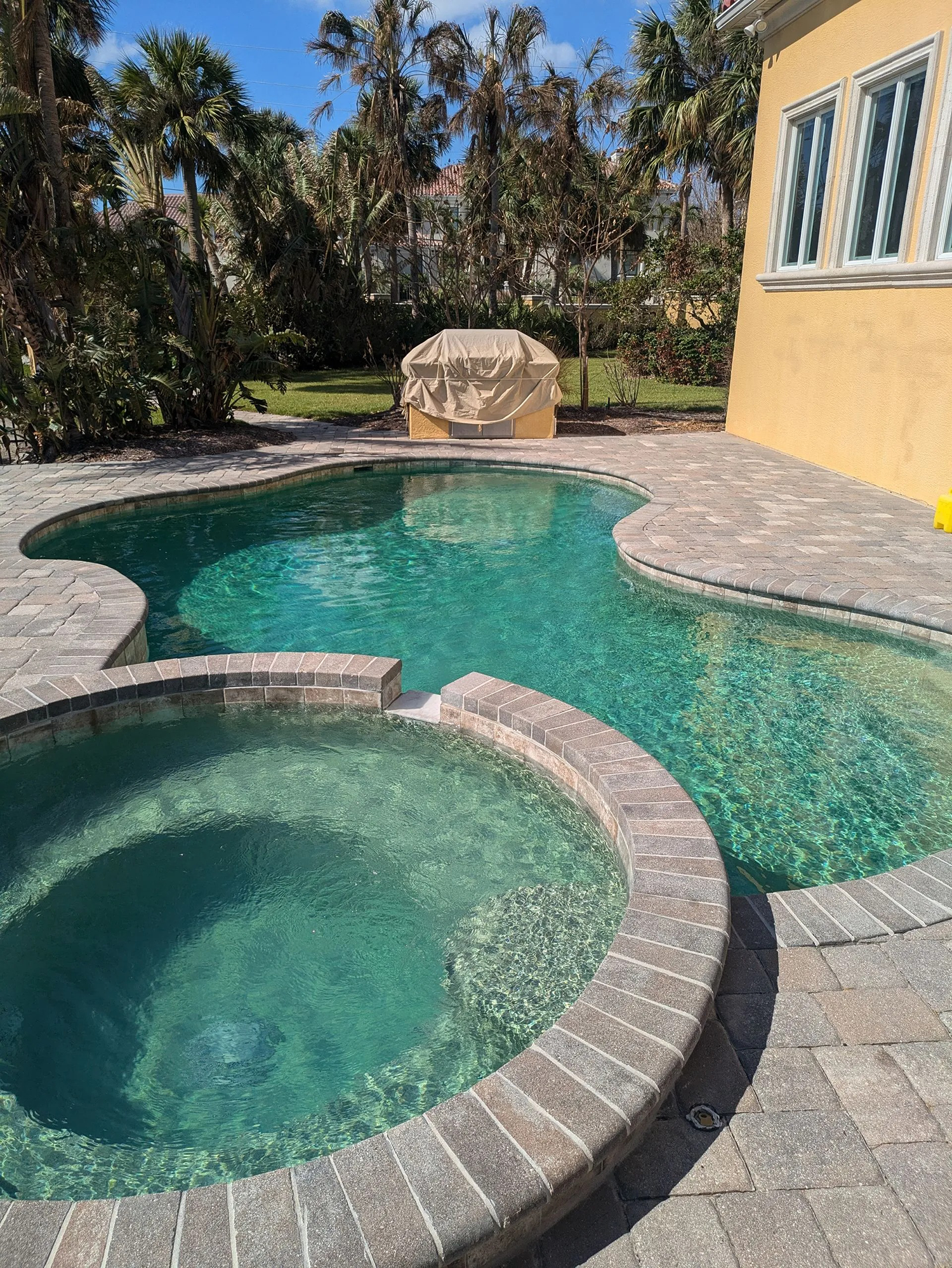 Swimming pool and spa with brick border, surrounded by patio, next to a yellow stucco building.