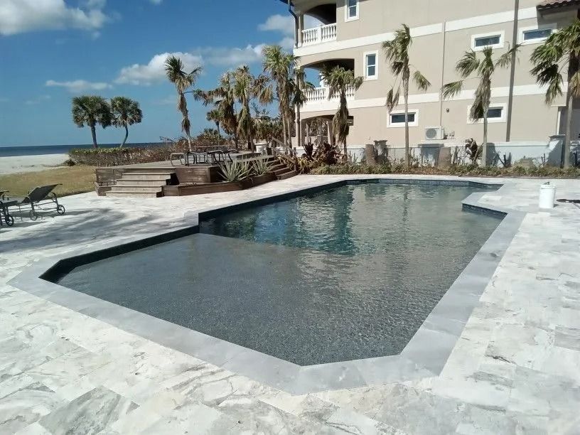 Pool overlooking a beach, with palm trees and a light-colored building in the background.