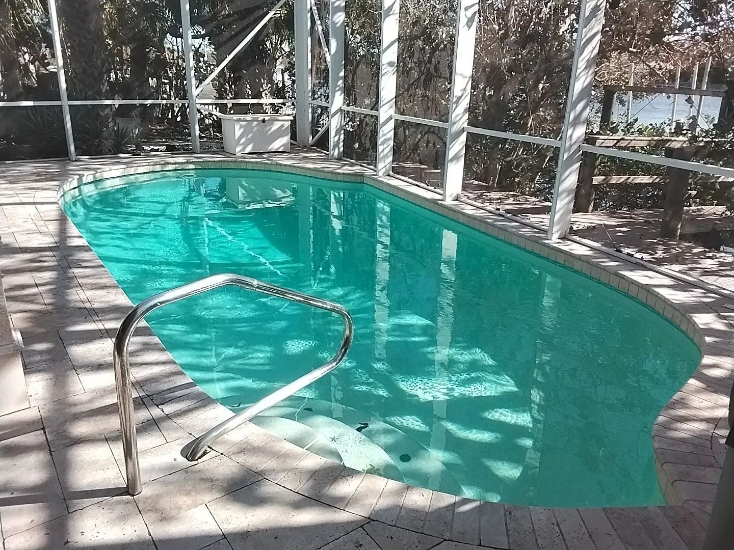 Pool surrounded by screen enclosure and brick pavers; teal water, metal handrail.