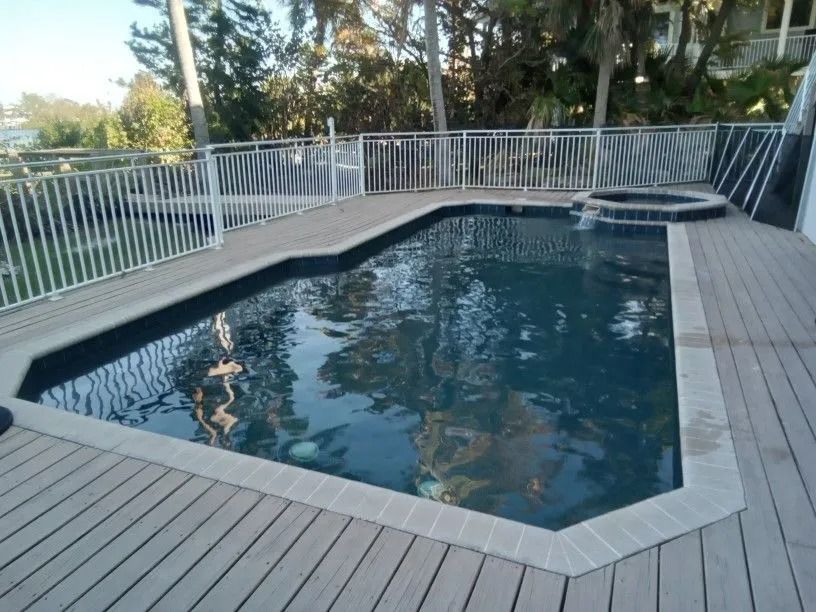 Pool with gray decking, white railing, and a hot tub. Trees in the background.