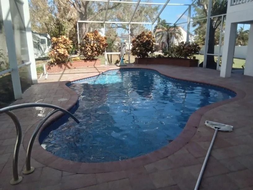 Pool surrounded by brick patio, enclosed by a screen. Blue tiled pool with water. Palm trees in the background.