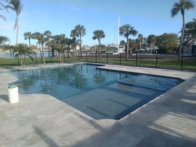 Swimming pool with steps, surrounded by light concrete. Black fence, palm trees, and waterfront in the background.