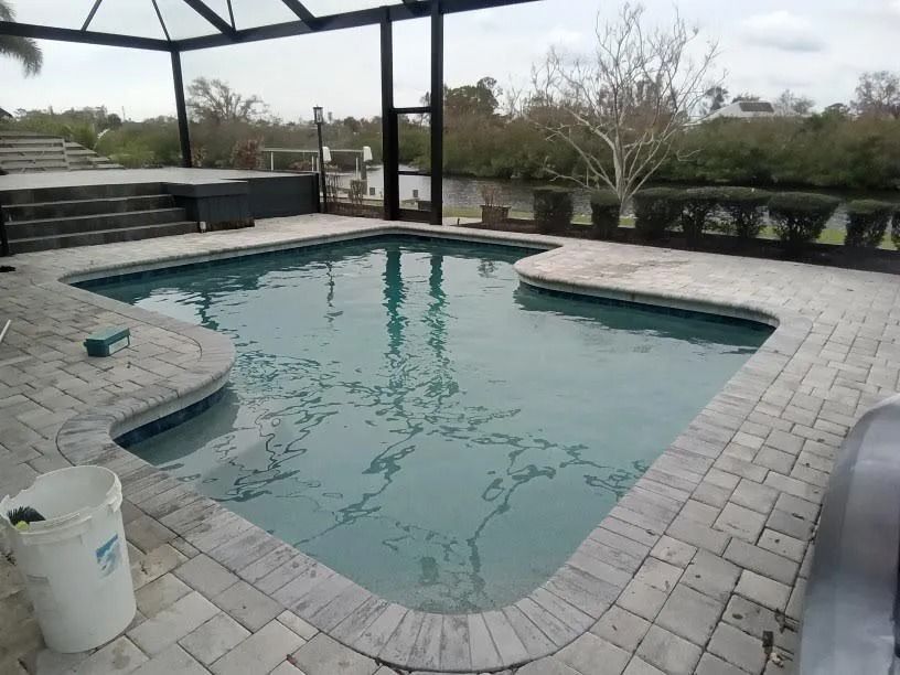 Pool with light-colored brickwork, under a screened enclosure. Lush greenery in the background, likely waterfront.
