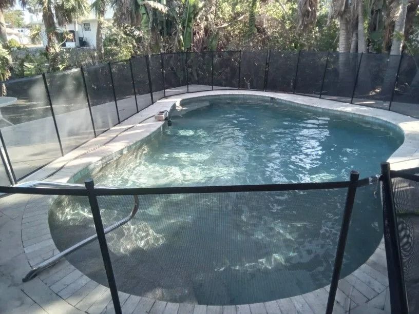 A pool with a black mesh safety fence. The water is light blue.  Surrounding is brick patio and trees.