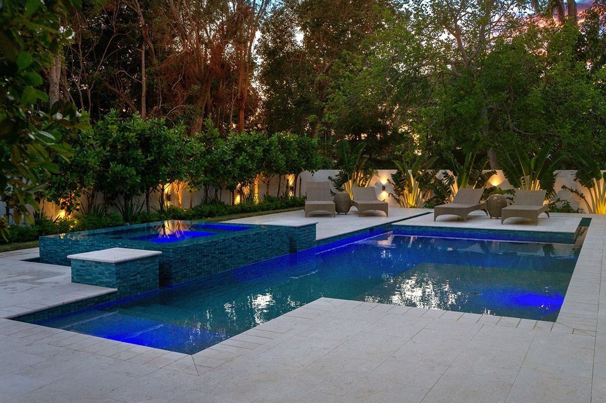 Pool and jacuzzi with blue lights surrounded by lounge chairs and lush greenery.