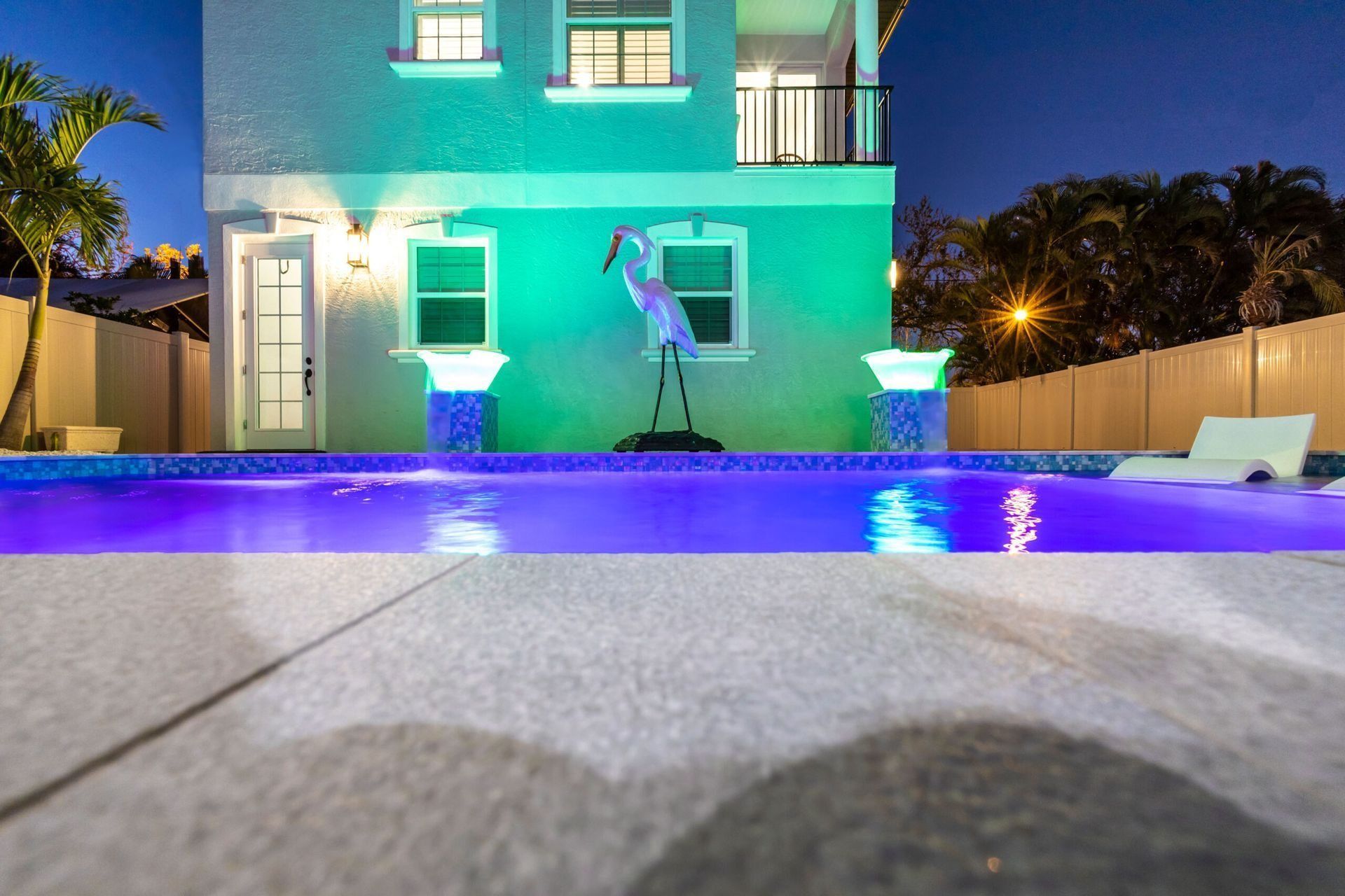Pool at night, lit with blue lights, in front of a modern house, a heron statue is centered.